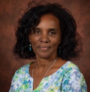 Portrait of a smiling woman with short curly black hair, turquoise earrings, wearing a blue-green floral blouse against a brown studio backdrop.