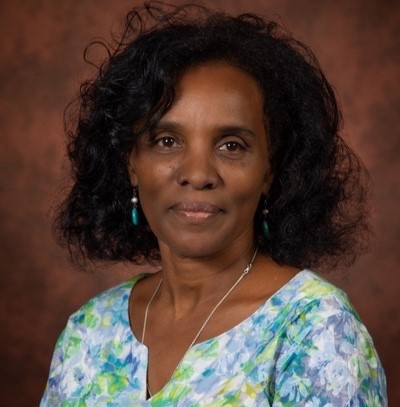 Portrait of a smiling woman with short curly black hair, turquoise earrings, wearing a blue-green floral blouse against a brown studio backdrop.