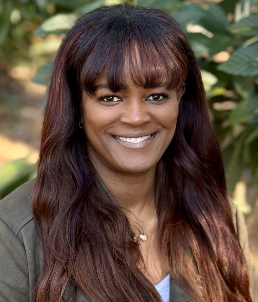 Close-up portrait of a smiling woman with long dark maroon hair and bangs outdoors.