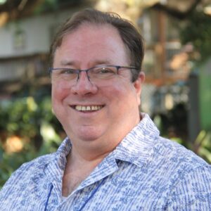 Close-up of a smiling man wearing blue-rimmed glasses, outdoors with blurred greenery in the background.